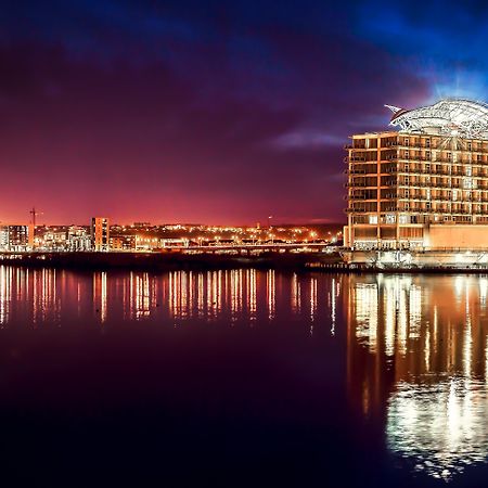 Voco St. David'S Cardiff By Ihg Hotel Exterior photo The photo depicts a modern building, likely a hotel or office complex, situated by a waterfront. It features a distinctive architectural design with a curved roof and multiple levels of glass windows. The scene is captured during twilight, creating a